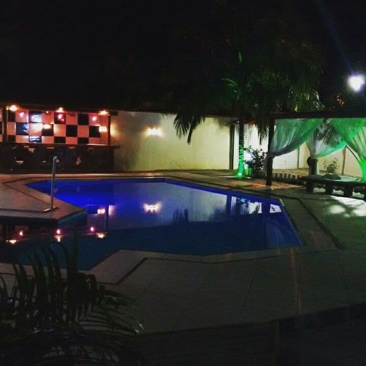 A well-lit pool area is featured, with water reflecting cool blue hues. Surrounding palm trees add a tropical touch. A gazebo with sheer drapes offers a shaded seating space, while the bar area is illuminated with soft lighting, enhancing the outdoor ambiance.