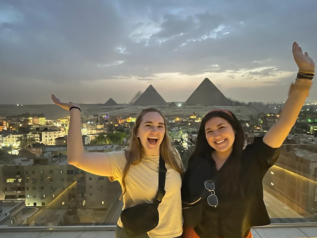 Two individuals stand with raised arms on a rooftop balcony, showcasing the majestic pyramids against a twilight sky. The cityscape below reflects warm lights, creating a contrasting backdrop to the monumental view. The scene captures a moment of camaraderie in a vibrant setting.