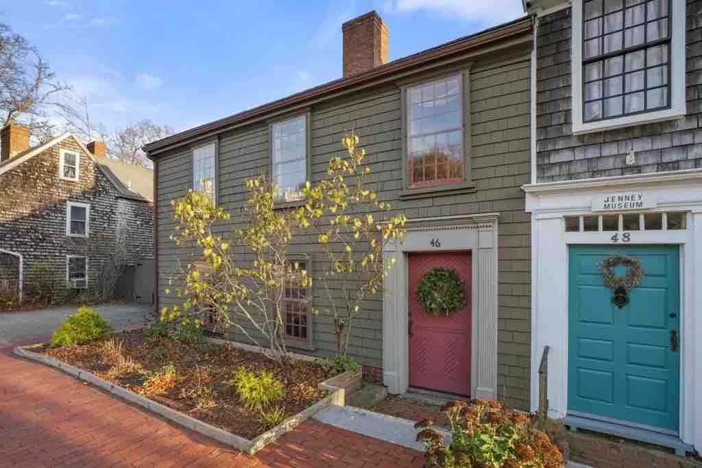 The image showcases the exterior of the historic Nathaniel Church House, revealing its weathered wooden siding and charming front door adorned with a wreath. A small garden is visible in the foreground, enhancing the inviting appeal of the property.