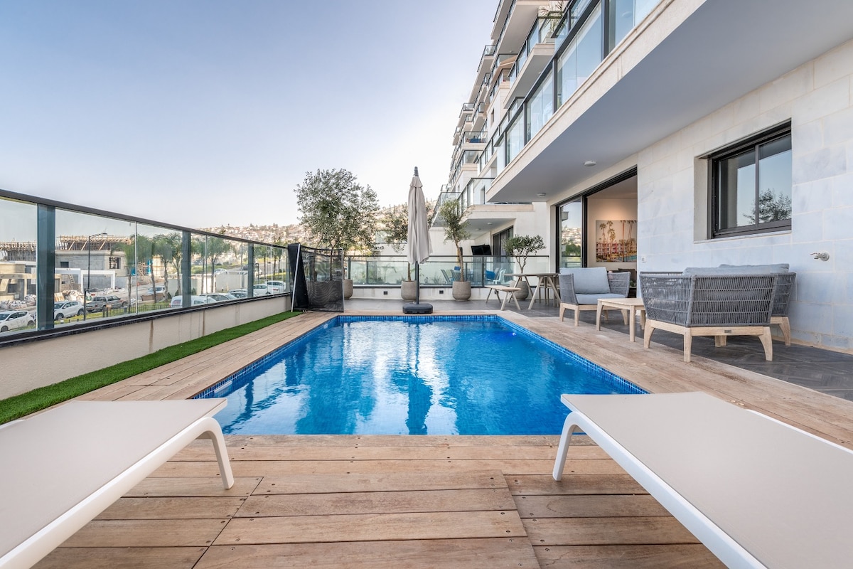 A private patio features a rectangular swimming pool surrounded by wooden decking. Two lounge chairs are positioned by the poolside, creating a relaxed atmosphere. The area is framed by glass railings, offering views of the surroundings, with a parasol providing shade.