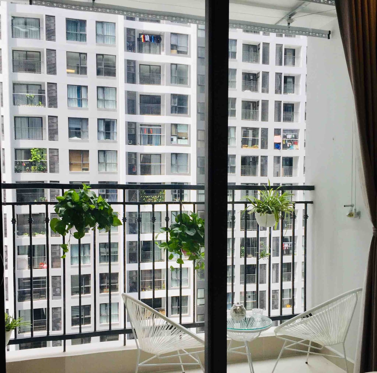 A balcony view presents a modern apartment complex in the background, featuring multiple windows. Two white chairs and a small round table are positioned on the balcony, accompanied by hanging plants that add a touch of greenery.