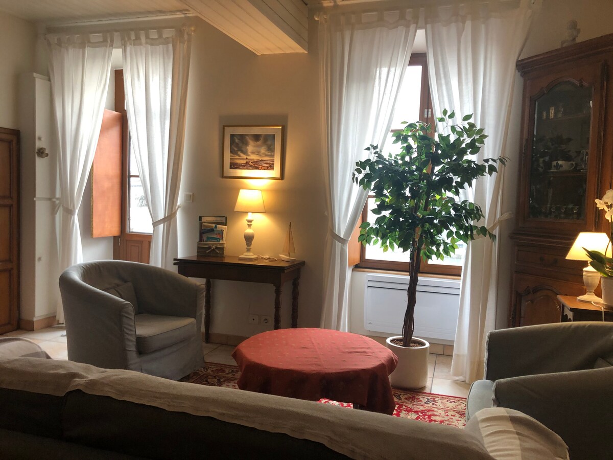 A cozy sitting area features two armchairs and a circular table, surrounded by natural light from large windows with white drapes. A small indoor plant adds a touch of greenery, complementing the warm lighting from nearby lamps and the wooden furniture present.