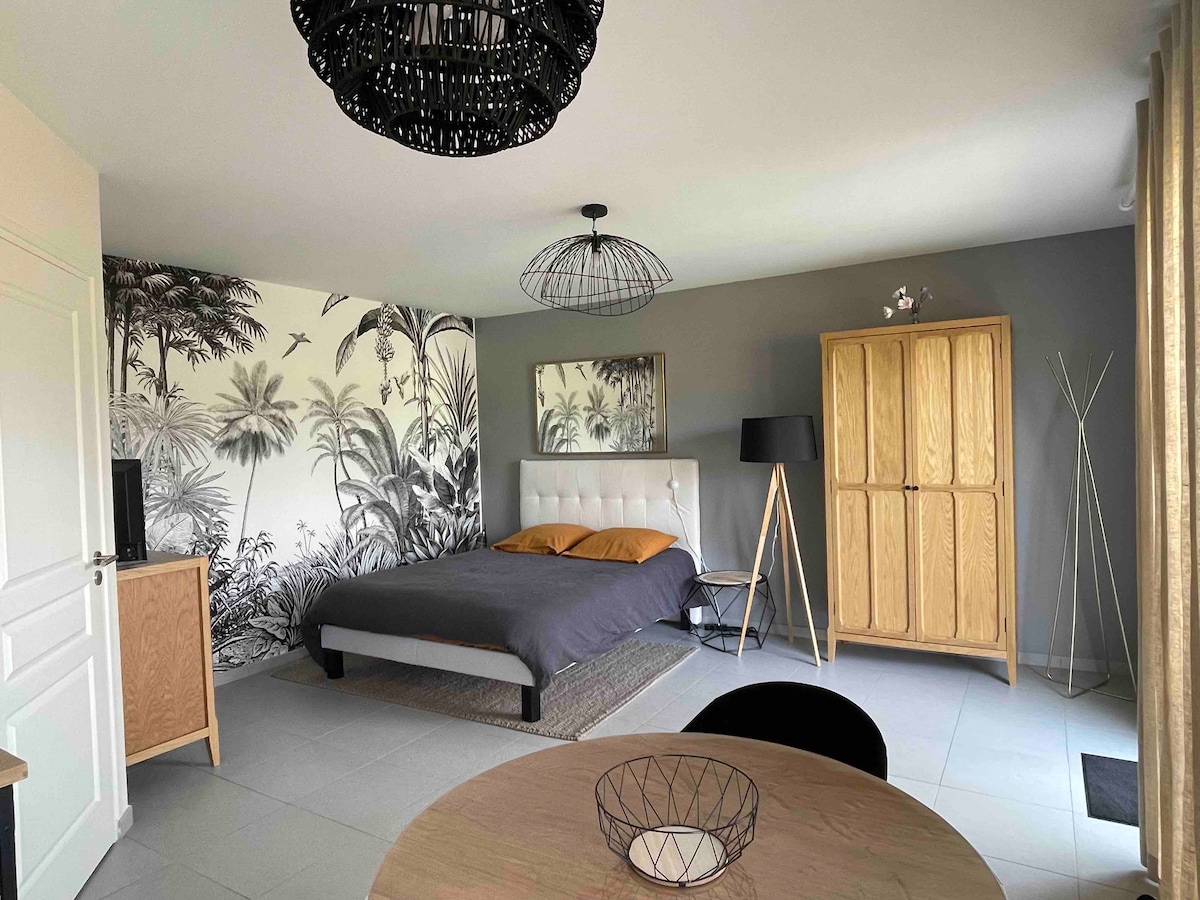 A spacious studio is highlighted by a large bed with neutral bedding and accented by decorative pillows. A stylish light fixture hangs above, and a modern lamp stands beside a wooden wardrobe. A round dining table is visible, contributing to the functional living space.