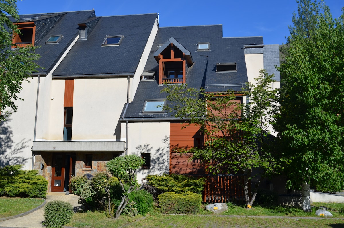 The apartment building features a modern design with a combination of stone and wood elements. Multiple windows and a gabled roof enhance its character. Surrounding greenery adds a touch of nature, while a pathway leads to the entrance, inviting guests to explore further.