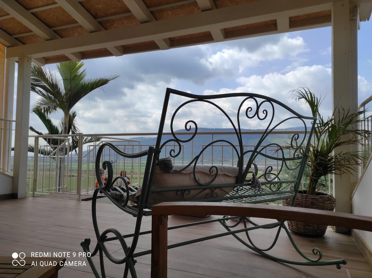 A furnished balcony features a wrought iron seating area, providing views of the surrounding landscape. Lush greenery and a palm tree are visible in the background, enhancing the tranquil setting. The sky is partially cloudy, creating a serene atmosphere.