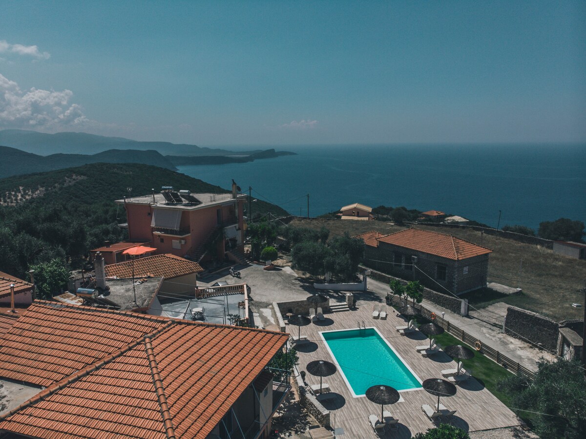 An aerial view presents a serene landscape featuring a well-maintained outdoor pool surrounded by lounge chairs and umbrellas. The rooftops of nearby buildings are visible, with distant hills and the sparkling blue sea in the background, creating a tranquil coastal atmosphere.