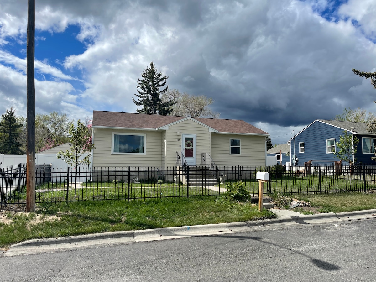 A single-story house is positioned at the end of a quiet street, featuring a light-colored exterior and a gently sloping front yard. A fenced area surrounds the property, with steps leading to the front door. Cloudy skies create a calm ambience overhead.