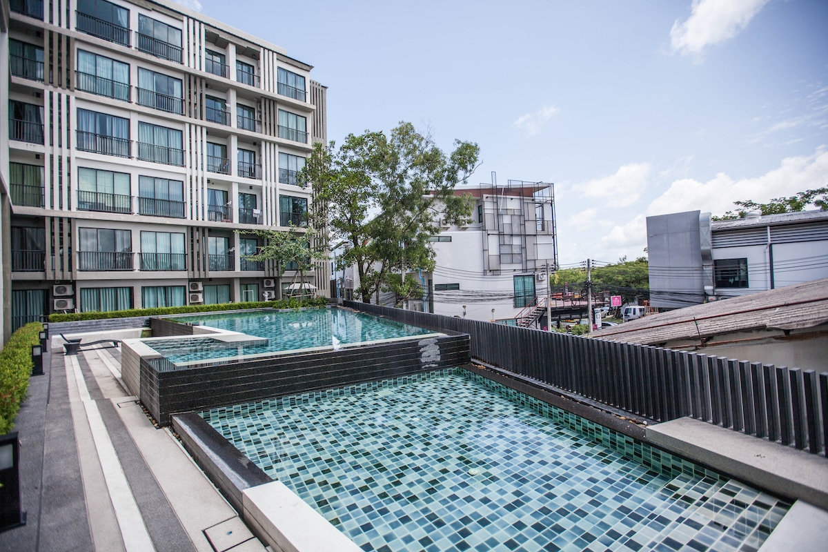 The image features an outdoor swimming pool area surrounded by modern apartment buildings. Two distinct pools, one for adults and one for children, are presented with mosaic tiles in shades of blue and green. The area is enhanced with greenery and a clear sky overhead.