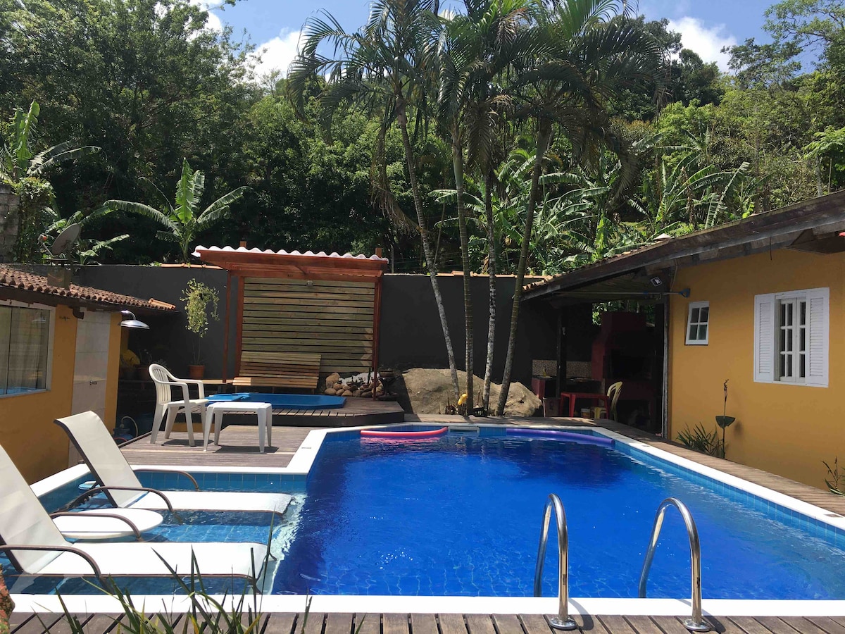 A refreshing swimming pool is surrounded by a wooden deck, featuring two lounge chairs and a small table. Lush greenery frames the area, complemented by a nearby rock formation. Colorful buildings with white-framed windows provide a vibrant backdrop.