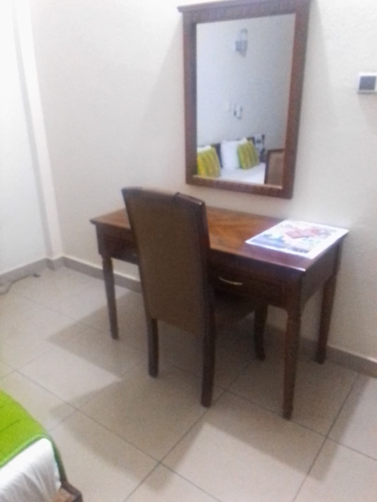 A wooden desk is positioned against a wall, accompanied by a chair. Above the desk, a mirror reflects the space. Natural light enters through a nearby window, while the floor is covered with tiled flooring, providing a clean and organized environment.