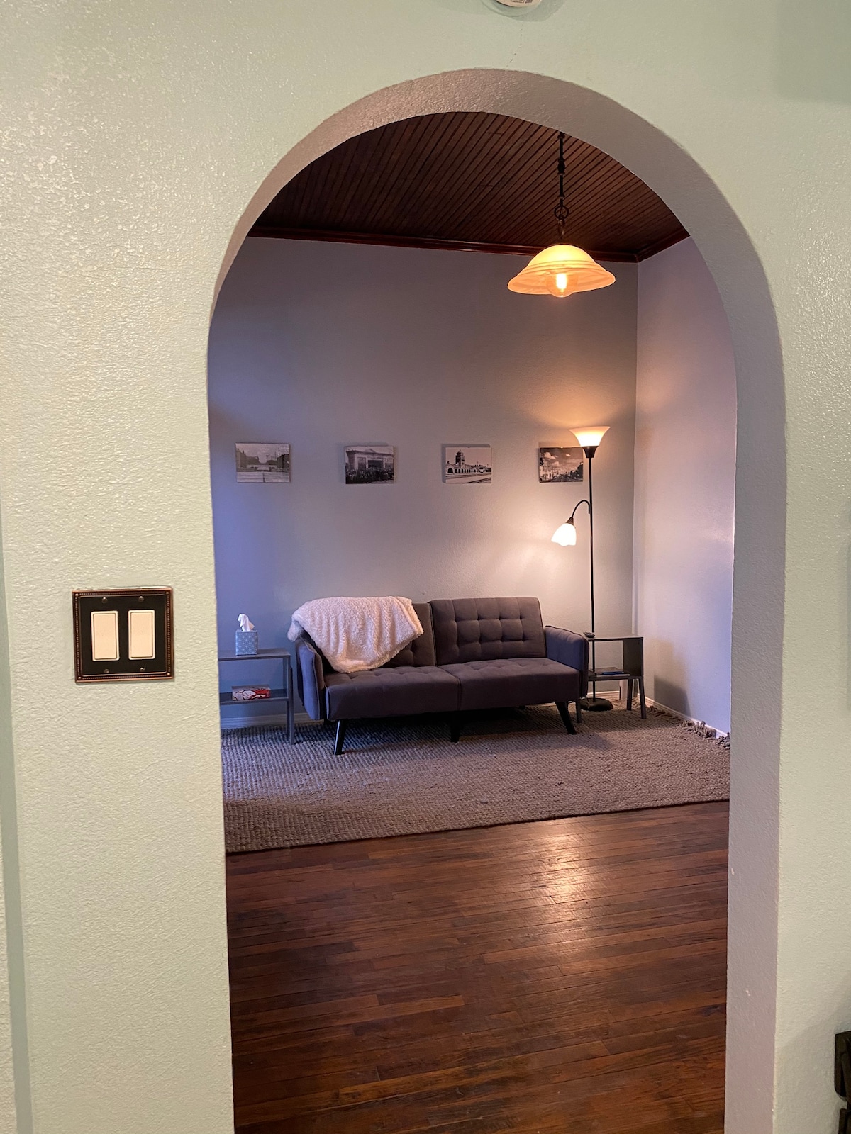 An inviting living area is seen through an arched doorway. A modern gray couch is positioned on a textured rug, accompanied by a floor lamp and a side table. Black-and-white photographs adorn the walls, and a soft blanket is laid across the couch.