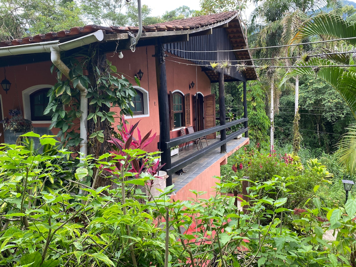 A charming house is surrounded by lush greenery, showcasing vibrant plants and flowers. The wooden balcony with black railing provides a scenic view of the natural landscape. The exterior features a warm pink hue, complemented by dark wooden details, creating a welcoming presence in the rural setting.