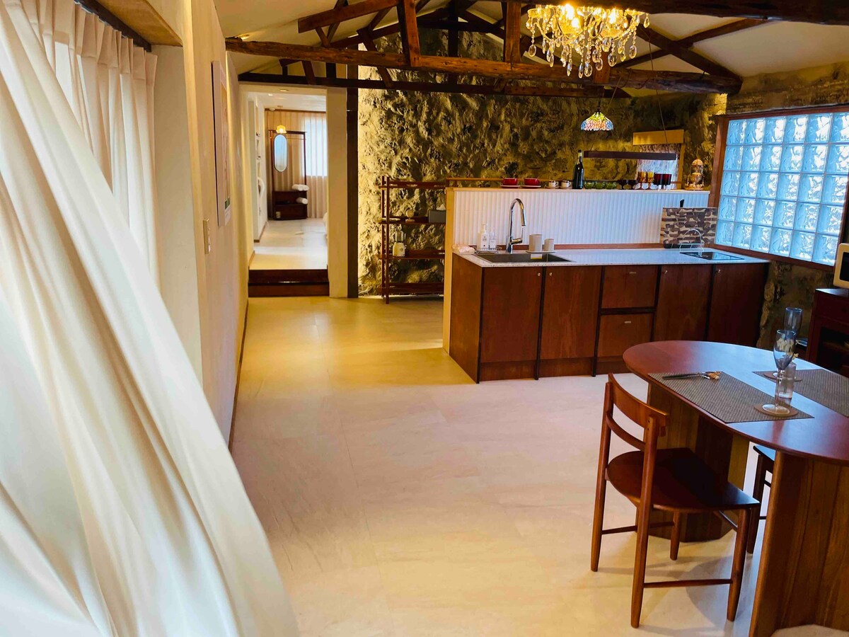 An open interior space features natural stone walls and wooden beams. A modern kitchen area is complemented by a wooden dining table and chairs. Soft lighting from a chandelier bathes the room, while large windows provide ample natural light.