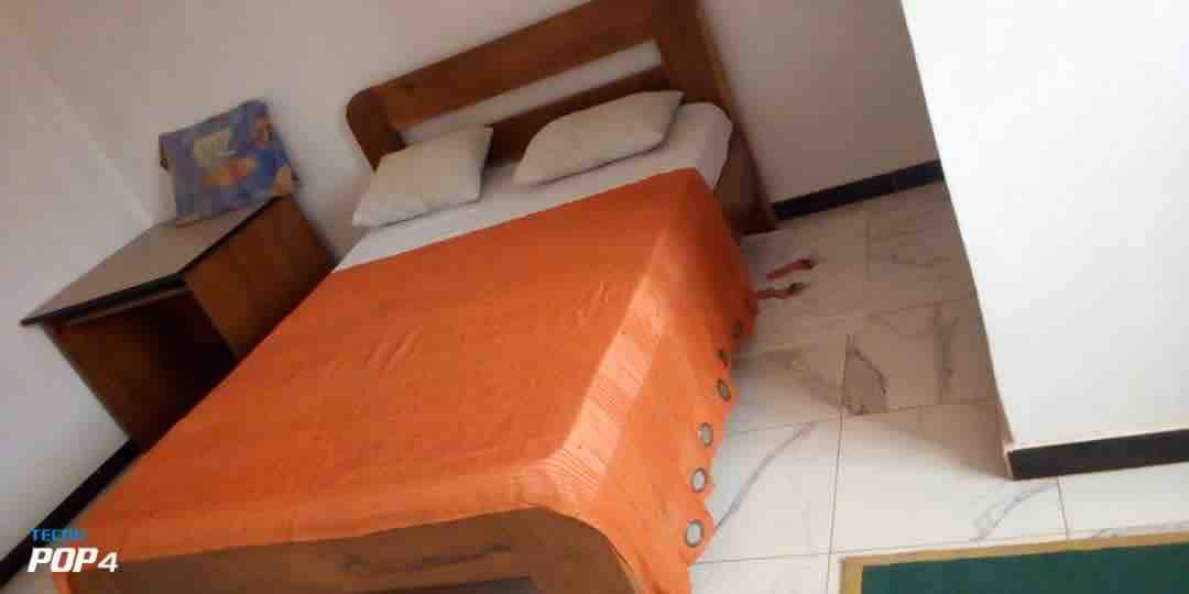 A modest bedroom features a spacious bed covered with an orange bedspread. Two pillows rest at the head of the bed. A wooden bedside table stands nearby, accompanied by a wall-mounted picture in the corner. The floor is tiled with light-colored, marbled patterns.
