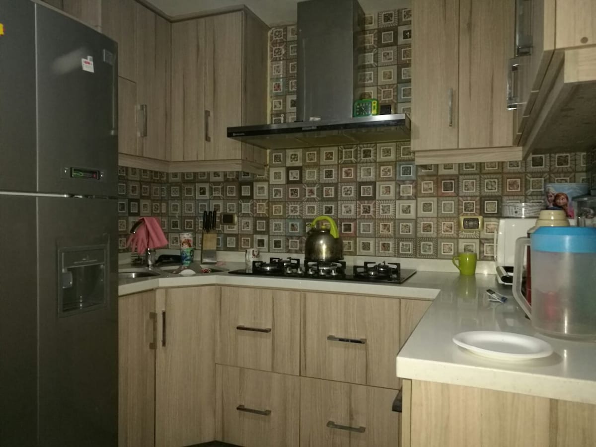 The kitchen features modern cabinetry with a mix of wood tones and intricate tiled backsplash. A stove is positioned on the counter, accompanied by utensils and appliances. A refrigerator stands on one side, creating a functional cooking space that is well-organized and inviting.