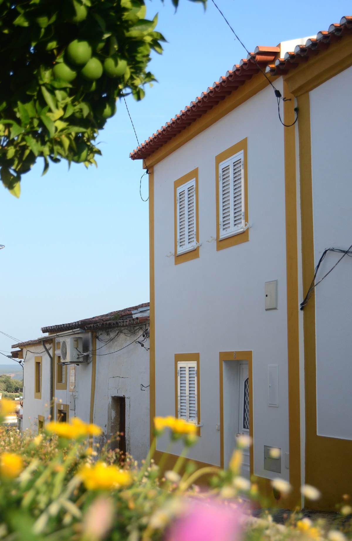 The exterior of a two-story house is captured, featuring a white façade with bright yellow details around the windows. Flowers in the foreground add color, while a clear blue sky enhances the setting. Nearby, another building with an unpainted exterior is visible.