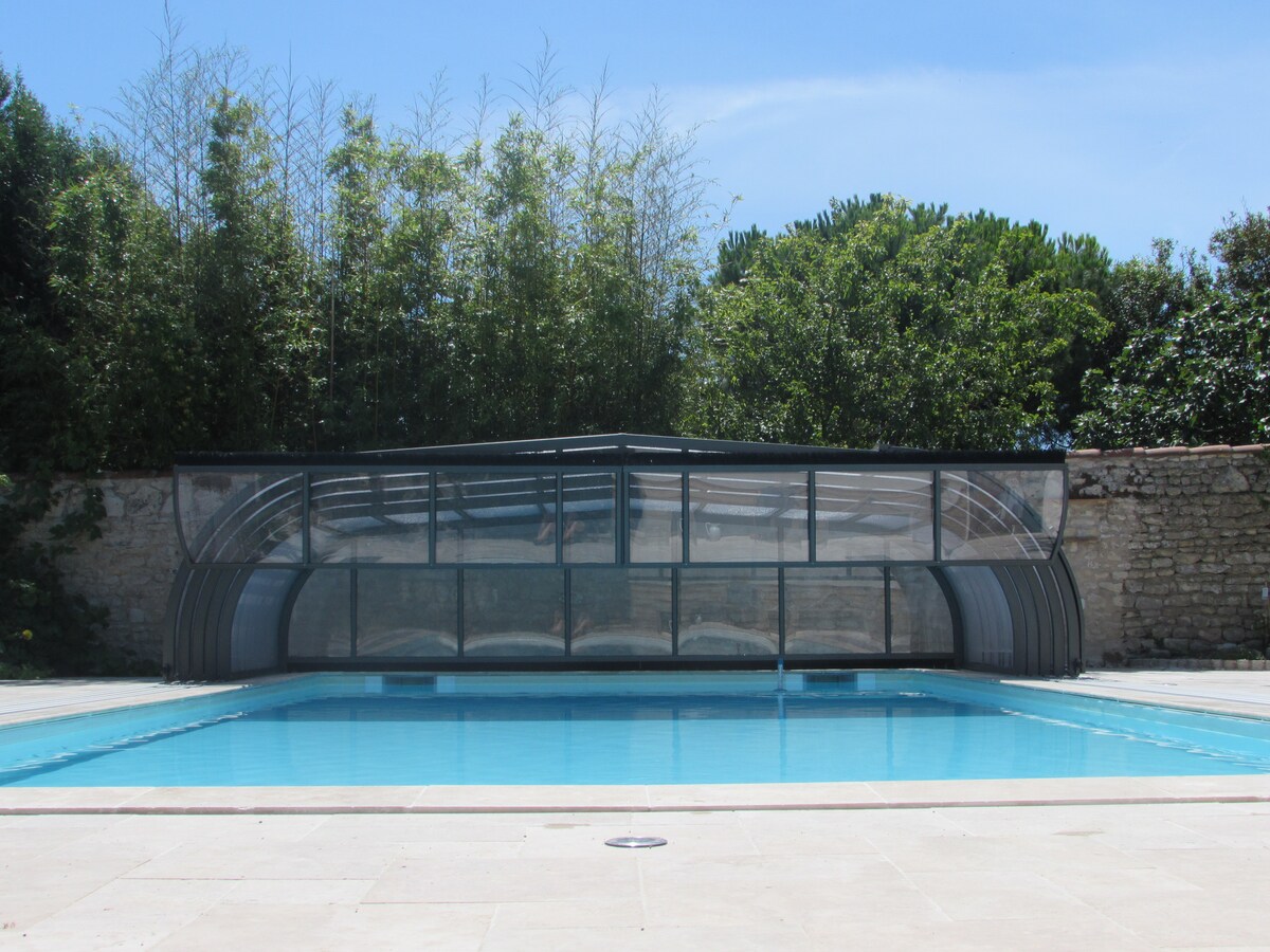 A collective heated pool is showcased, featuring a modern protective cover. The pool is surrounded by paved stone and lush greenery, including tall foliage and trees, providing a serene backdrop. Clear blue water reflects the sky, creating a tranquil setting for relaxation.