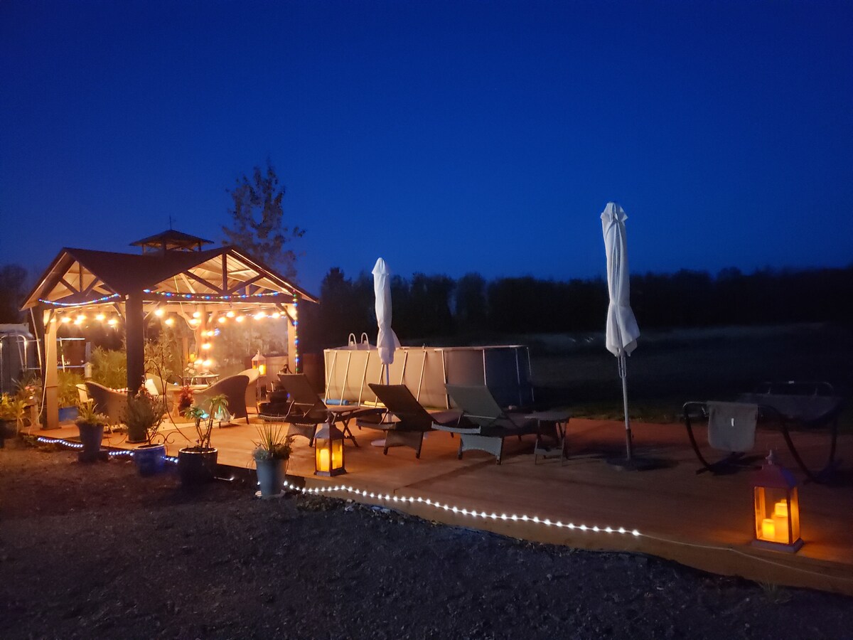 An outdoor gazebo is illuminated by string lights, surrounded by comfortable sun beds and lanterns. The wooden deck extends into an open area with low vegetation, offering a serene ambiance at night. Nearby, umbrellas are positioned to provide shade during the day.
