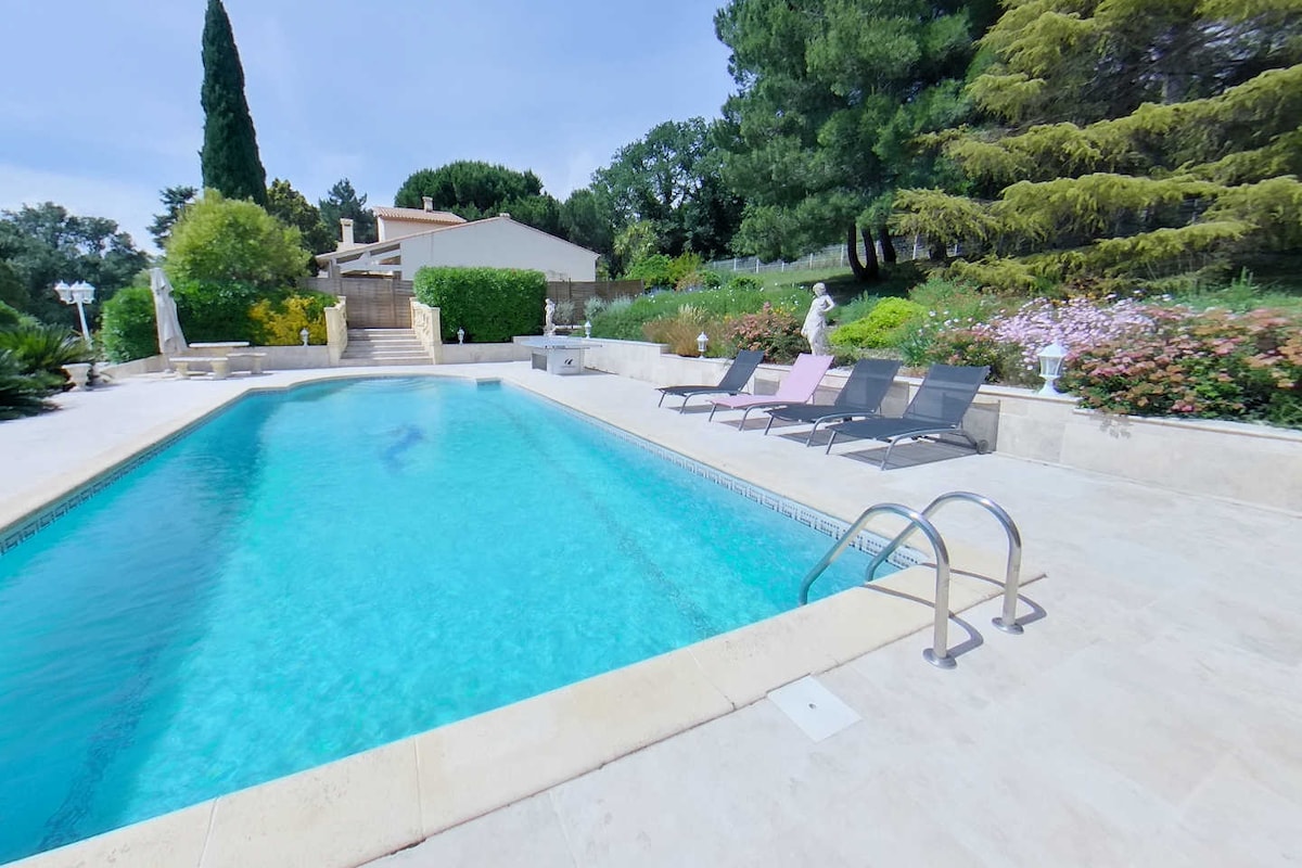 A serene outdoor area features a swimming pool, surrounded by well-kept gardens filled with blooming plants. Five sun loungers are positioned beside the pool, and steps lead to an adjacent terraced area. Lush trees provide shade, enhancing the peaceful ambiance.