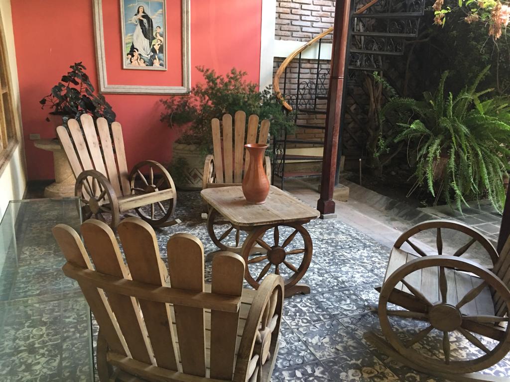 A serene interior garden area features four wooden chairs designed with a wagon wheel motif, arranged around a rustic table. A vase rests in the center, surrounded by lush greenery and indoor plants, while a spiral staircase is visible in the background.