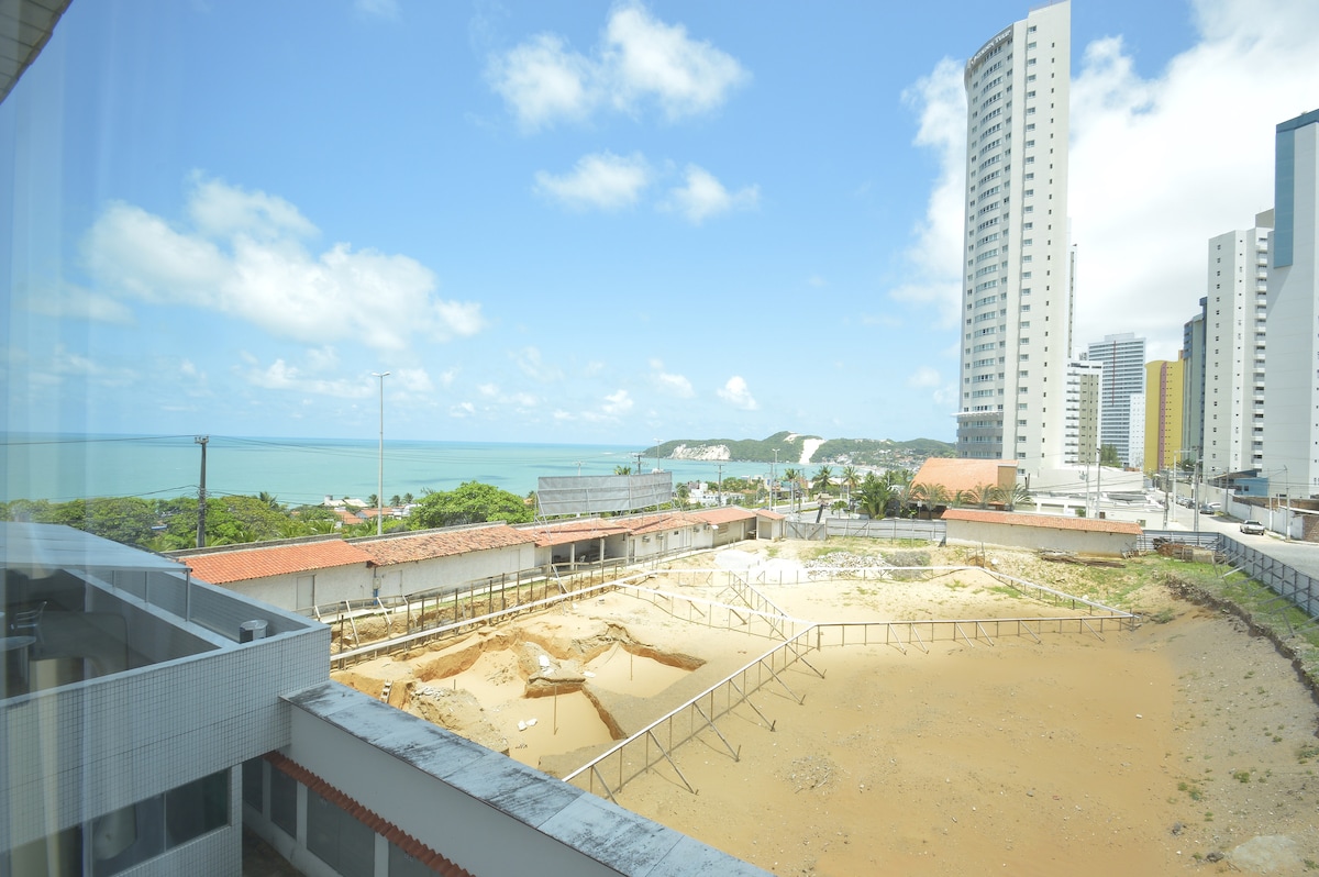 A view from the apartment showcases a clear sky with soft clouds above an area of sand and earth below. To the left, the coastline can be seen, and distant hills rise towards the right, creating a serene backdrop against modern buildings.