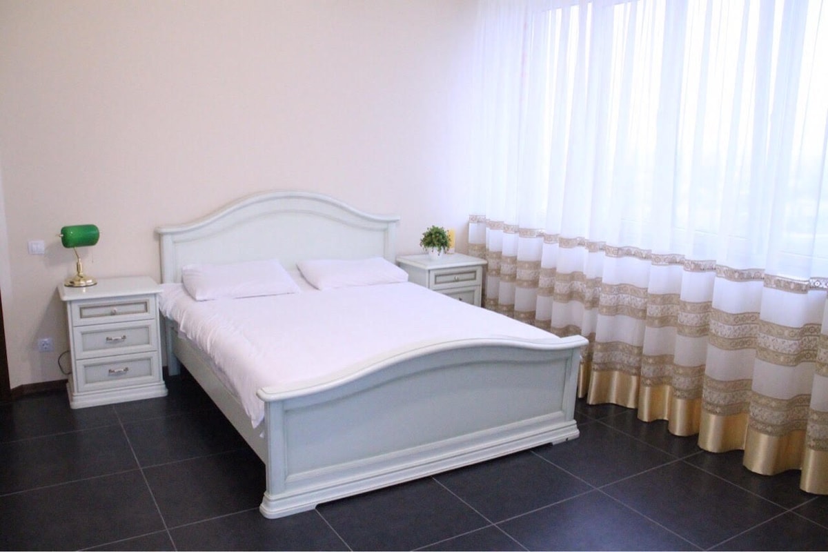 A bright sleeping area is highlighted by a spacious bed with a light-colored frame and crisp white bedding. Two nightstands flank the bed, complemented by a green lamp. Natural light filters through the large window, adorned with sheer and patterned curtains.