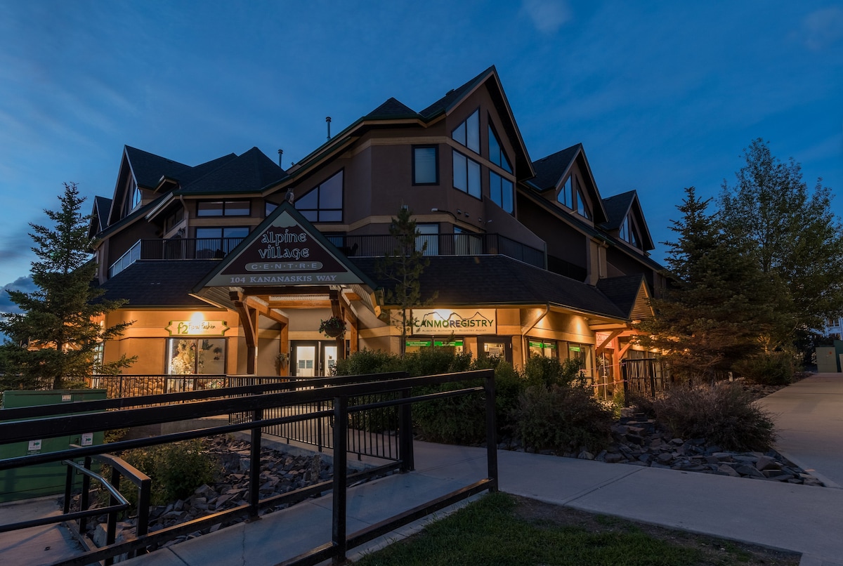 The exterior of the two-story condo complex is illuminated at dusk, showcasing a combination of natural and artificial lighting. Surrounding trees and shrubs enhance the landscape, while signage indicates nearby amenities. A pathway leads to the entrance, creating a welcoming access point.