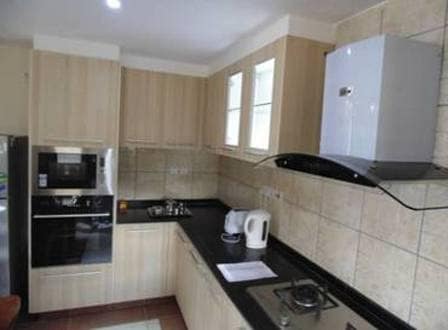 A modern kitchen features light wood cabinetry, a sleek black countertop, and stainless steel appliances. A gas stove with a hood and a built-in oven are visible. Natural light enters through windows above the sink, enhancing the functional layout of the space.