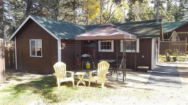 The exterior of the cottage showcases a brown wooden facade, complemented by a green roof. A shaded gazebo is positioned on the patio, accompanied by two yellow chairs and a small table. A barbecue grill is visible nearby, surrounded by trees.