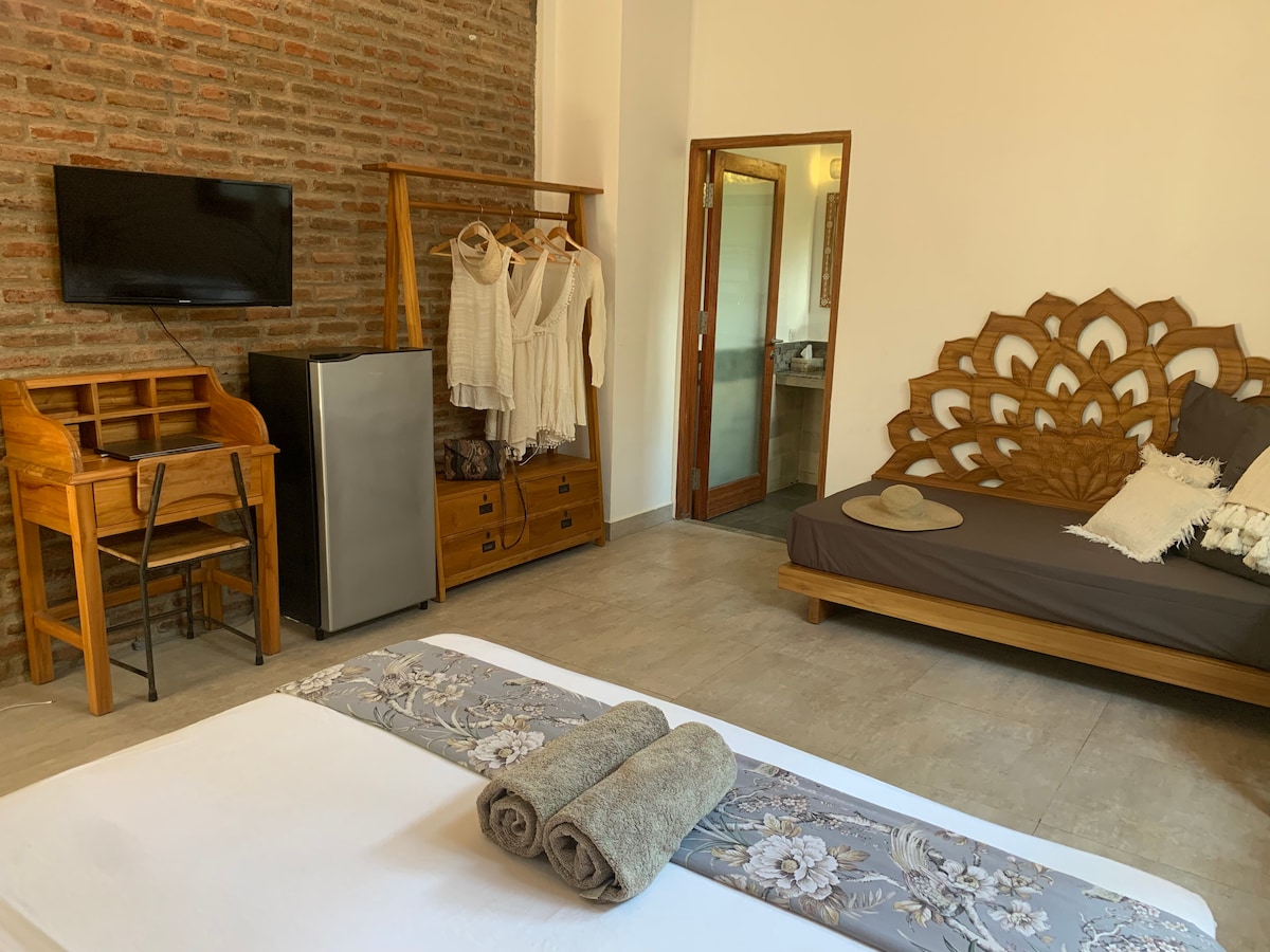 A spacious room featuring a king size bed and a sofa bed. A flat-screen TV is mounted on a brick wall, with a wooden garment rack and a small fridge positioned nearby. A desk with chairs offers a functional workspace, alongside a private bathroom visible through the open door.
