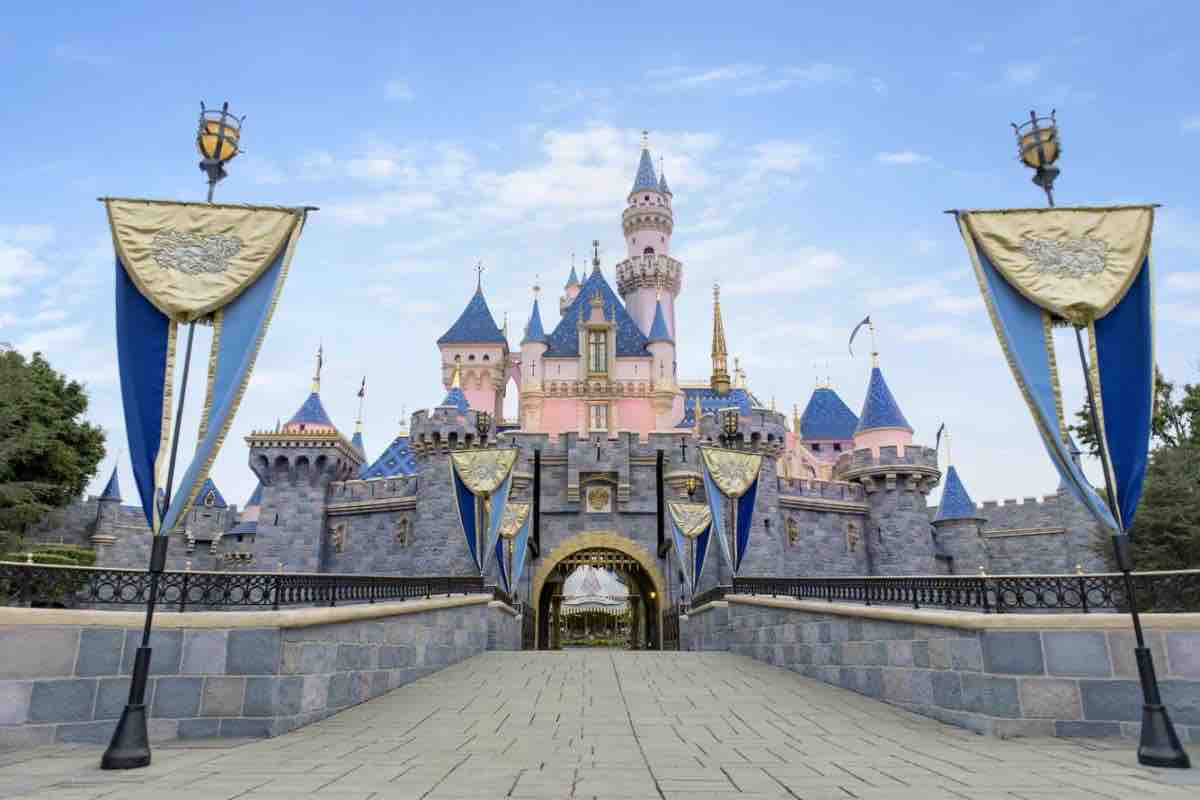 A castle with pink and blue spires is depicted against a clear sky. The entrance is flanked by decorative banners and lanterns, inviting visitors to explore the enchanting structure. A paved pathway leads towards the castle, providing a sense of approachability and wonder.