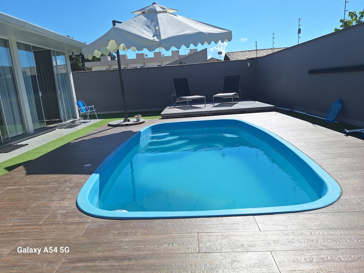 A private pool is framed by a wooden deck, surrounded by a shaded area featuring lounge chairs. A large umbrella provides shelter from the sun, while the pool's clear water reflects the bright blue sky.