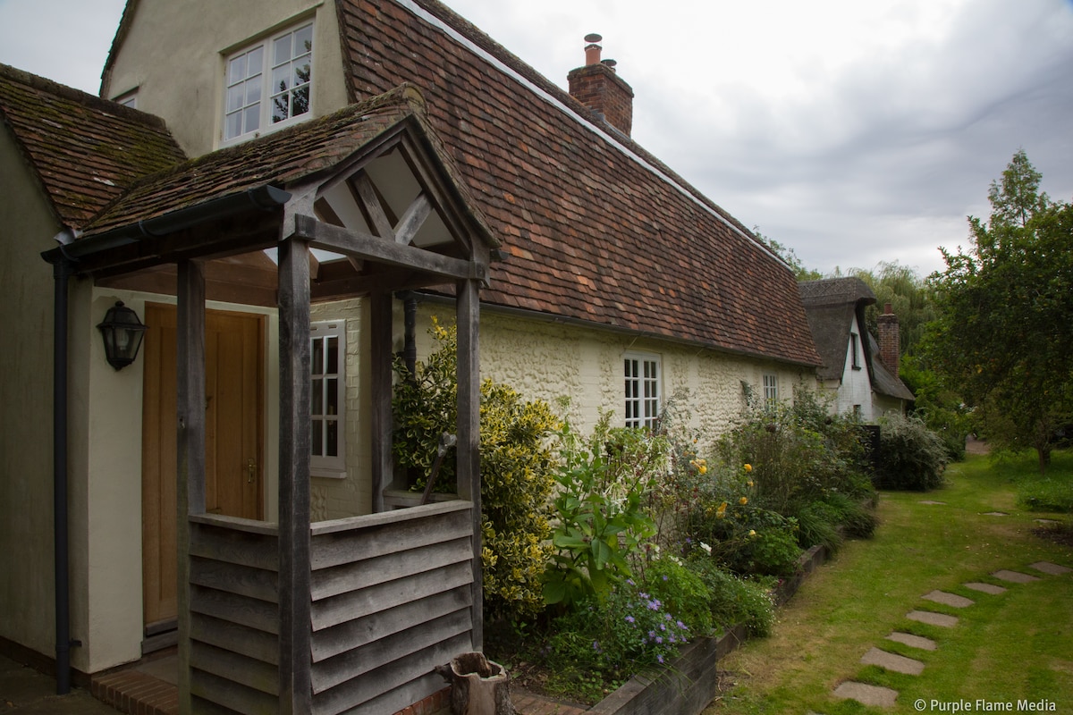 A charming cottage is presented, featuring a textured white exterior and a slate roof. A covered porch with wooden accents is visible in the foreground, while a pathway leads through well-maintained gardens with blooming flowers on either side.