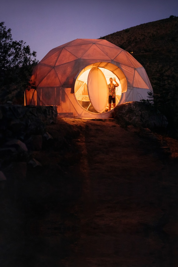 黃昏時分，一間造型獨特的圓頂屋沐浴在溫暖宜人的光線中，一位旅客站在門口揮手。