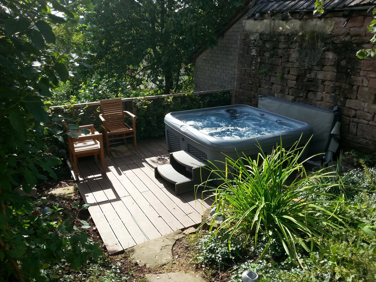 A private outdoor area features a hot tub surrounded by lush greenery. Wooden decking leads to the hot tub, with a comfortable chair positioned nearby. Natural light filters through the surrounding plants, creating a serene environment.