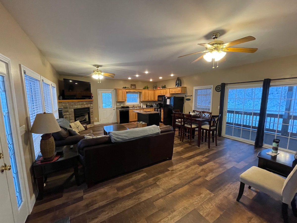 An open living area features a comfortable sectional sofa facing a fireplace and a dining table for six. The kitchen, visible in the background, includes wooden cabinetry and modern appliances. Large windows allow natural light, with sliding doors leading to the outdoor deck.