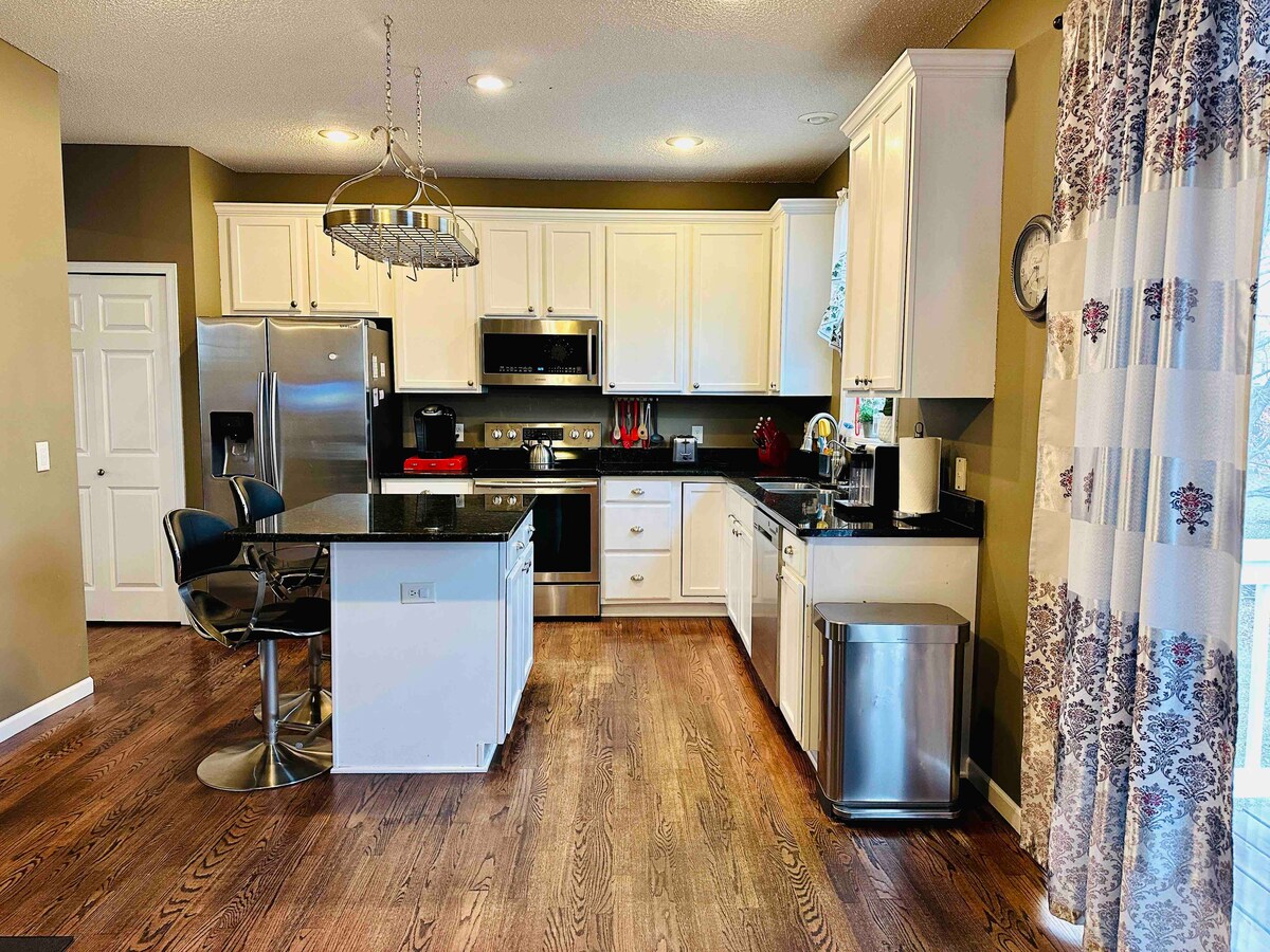 A modern kitchen is equipped with stainless steel appliances and white cabinetry. A central island with bar seating is featured, along with a dining area. Natural light enters through the window, highlighting the dark countertops and warm wooden flooring.