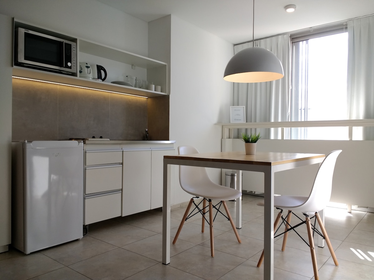 A bright kitchen area is shown, featuring modern white cabinetry and a small table with two chairs. A refrigerator and microwave are visible, along with a window that allows natural light to fill the space. A potted plant is placed on the table for a touch of greenery.