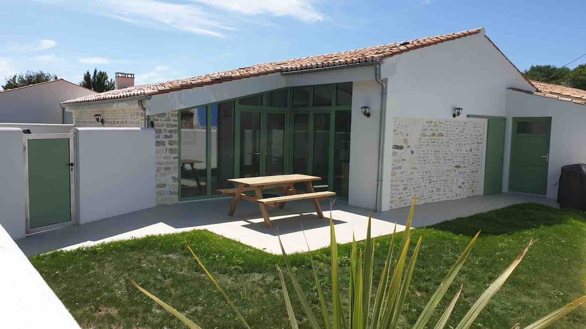 The exterior of a modern vacation home is shown, featuring a pitched roof and large glass windows. A wooden picnic table is positioned on a light concrete patio, surrounded by green grass and low landscaping. Two storage units are visible against the wall.