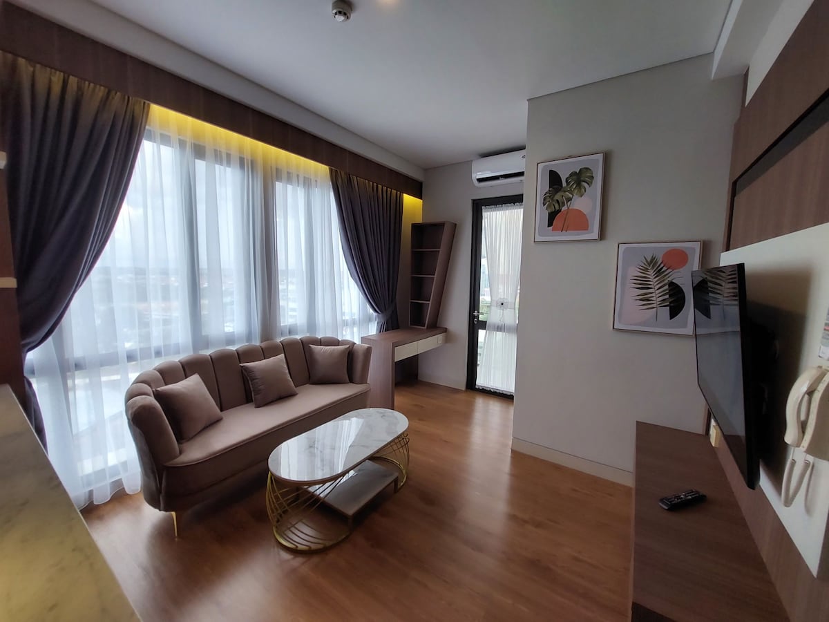 A contemporary living area features a plush sofa paired with a stylish coffee table. Large windows dressed in elegant curtains allow natural light to illuminate the space. Artwork depicting botanical designs adds a touch of style, while a wall-mounted television provides entertainment options.