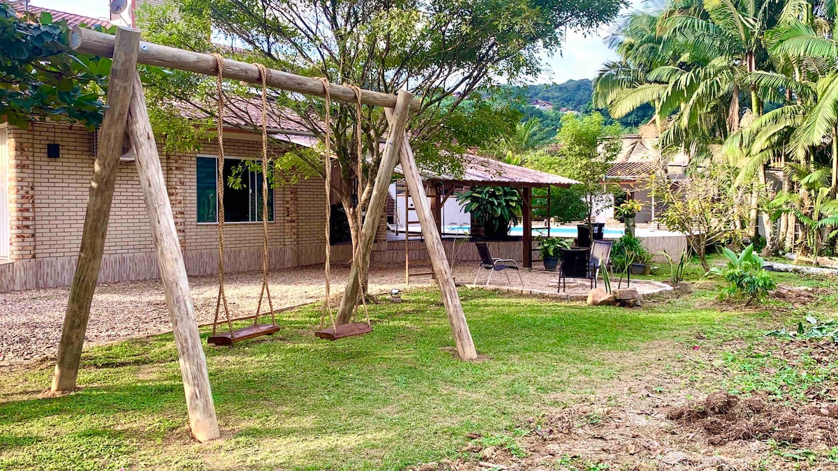 An outdoor area features a wooden swing set placed on a grassy lawn, surrounded by trees and plants. In the background, a shaded patio is visible, enhancing the space for relaxation and recreation.