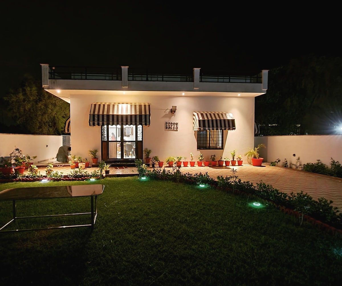 The exterior of the home is illuminated at night, featuring a well-kept garden with potted plants along the walkway. Large glass doors with striped awnings provide access to the interior. Soft lighting enhances the manicured lawn, creating a tranquil ambiance.