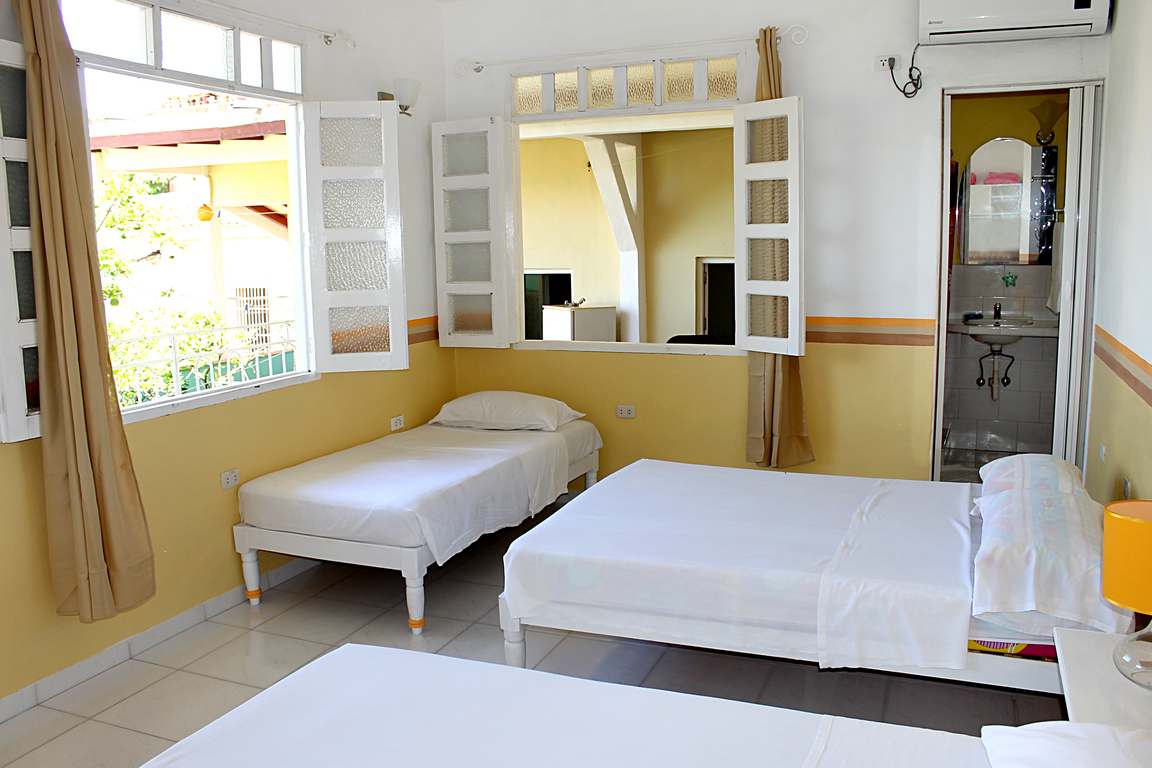A bright room features two spacious beds and a single bed, all dressed in crisp white linens. Large windows with wooden shutters allow natural light to fill the space, while a small bathroom is visible through a glass window in the corner.
