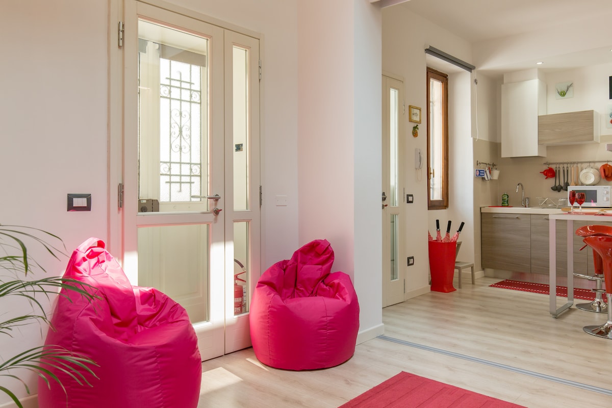 A bright entrance area features two pink bean bag chairs positioned beside glass doors. The interior flows into a modern kitchen area with light-colored cabinetry, and a small red mat adds a pop of color to the light wooden floor.