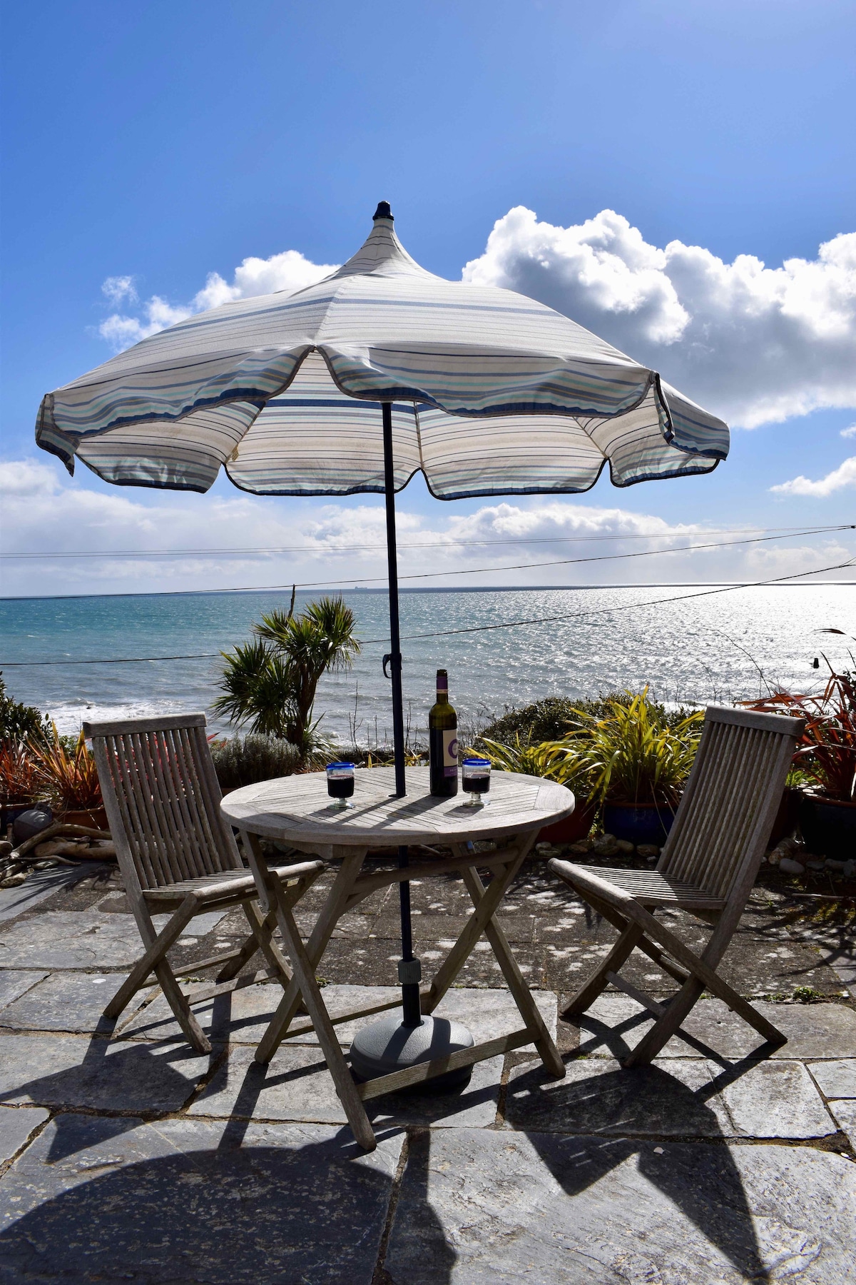 An outdoor dining area is situated on a slate patio, featuring a round wooden table with two chairs beneath a striped parasol. A bottle and glasses are placed on the table, with a backdrop of the sea and a clear blue sky, accented by scattered clouds.