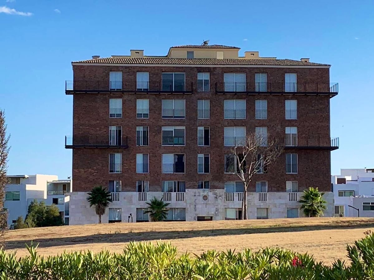 A modern brick building is depicted, featuring multiple balconies and large windows. The structure is surrounded by open grassy areas and palm trees, with clear blue sky above. The design incorporates a smooth stone base, providing a contrast to the textured brick facade.