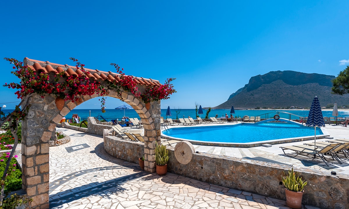 An inviting outdoor area features a large swimming pool surrounded by lounge chairs. A stone archway draped with vibrant flowers frames the view of the pool and the sea. The clear blue sky enhances the serene coastal atmosphere.