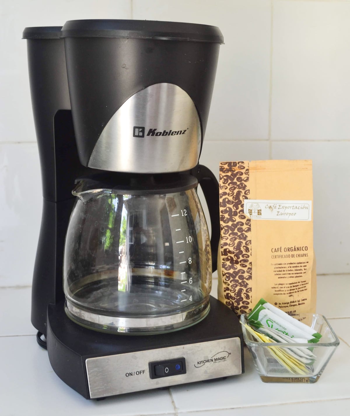 A coffee maker is positioned on a countertop, featuring a glass carafe with measurement markings. Next to it, a bag of organic coffee is visible, accompanied by a small dish containing coffee stirrers and a spoon. The setup invites a coffee-making experience.