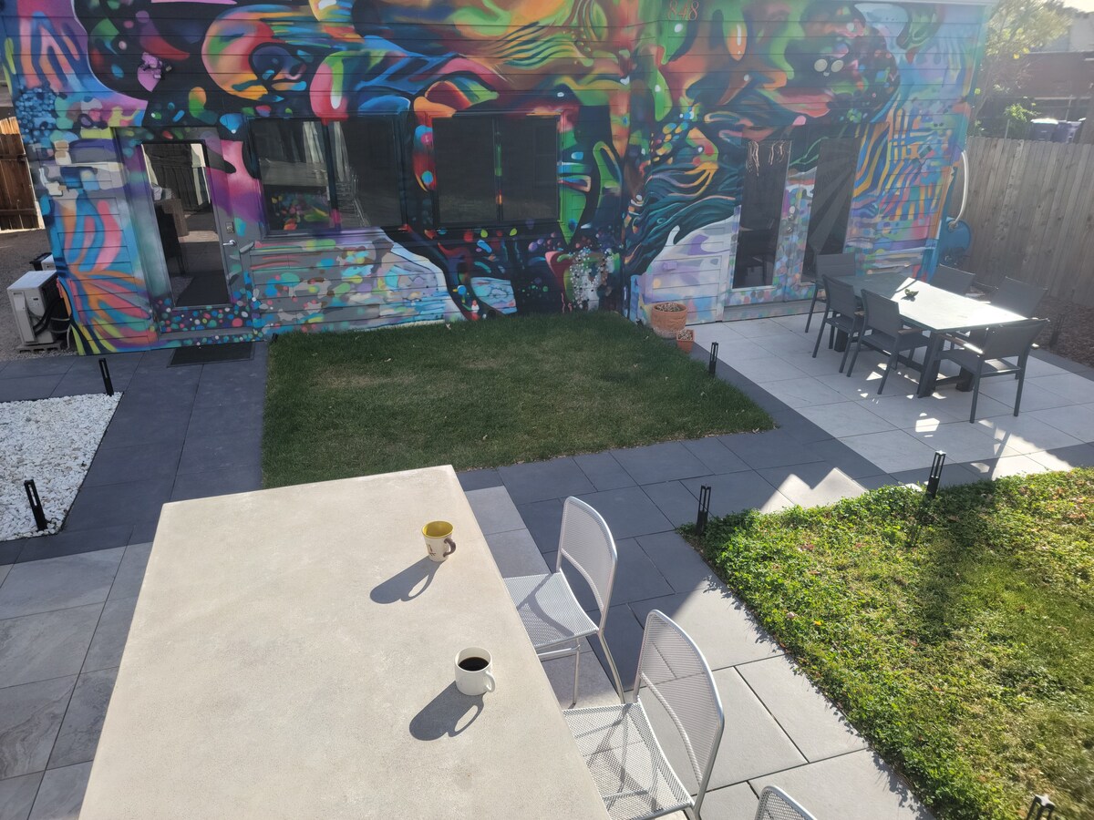 A vibrant outdoor space is showcased, featuring a colorful mural on the building's exterior. The patio has a dining area with dark furniture, while a grassy area complements the paved sections. Two chairs with cups are positioned on the stone tabletop, inviting relaxation.