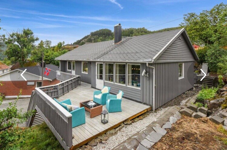 A gray-painted house features a spacious deck adorned with teal outdoor chairs surrounding a wooden table. Large windows provide ample natural light to the interior, while greenery and a hillside landscape create a serene backdrop.